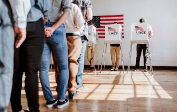 Interior, a line of voters while several people are standing behind voting booths with an American flag on the wall behind them